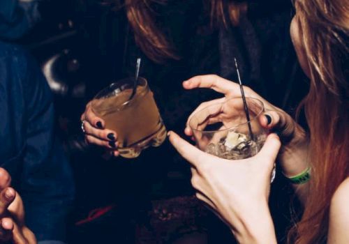 A group of people are socializing, holding drinks and chatting in a dimly lit setting. They appear to be enjoying themselves, laughing and having fun.