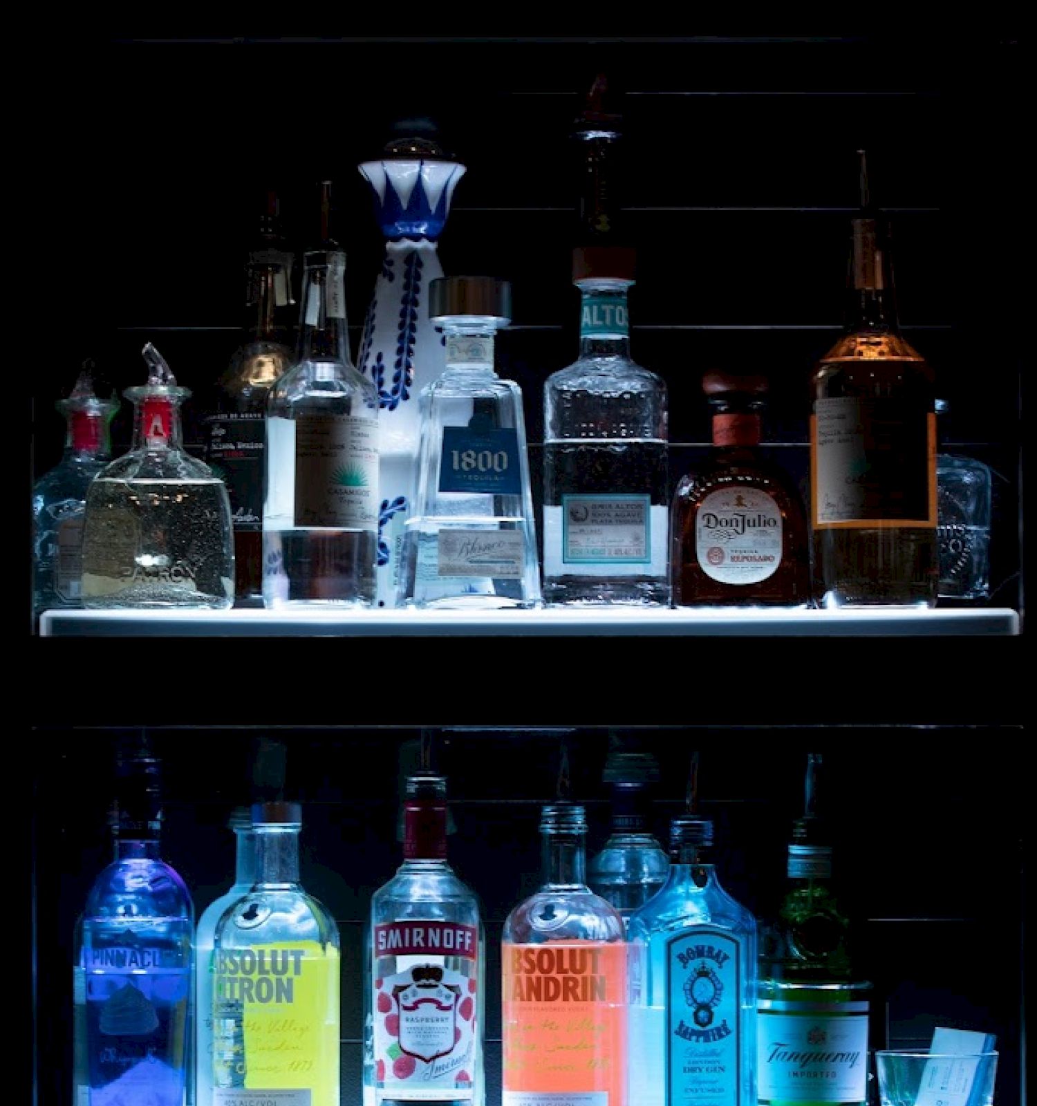 The image shows two shelves filled with illuminated bottles of various alcoholic beverages, including vodka, gin, whiskey, and tequila.