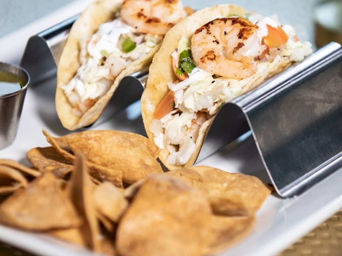 The image shows two tacos filled with ingredients like shrimp, served on a metal taco holder, with a side of chips on a white plate.