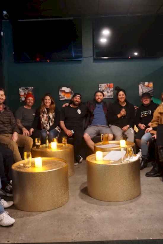 A group of people are sitting on black sofas around golden tables with candles, smiling at the camera in a cozy indoor setting.