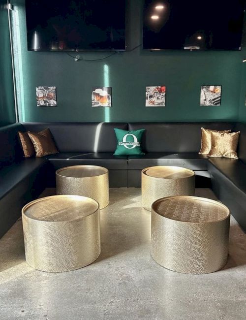 A modern lounge area with black couches, gold pillows, and round gold tables, decorated with framed pictures on green walls.