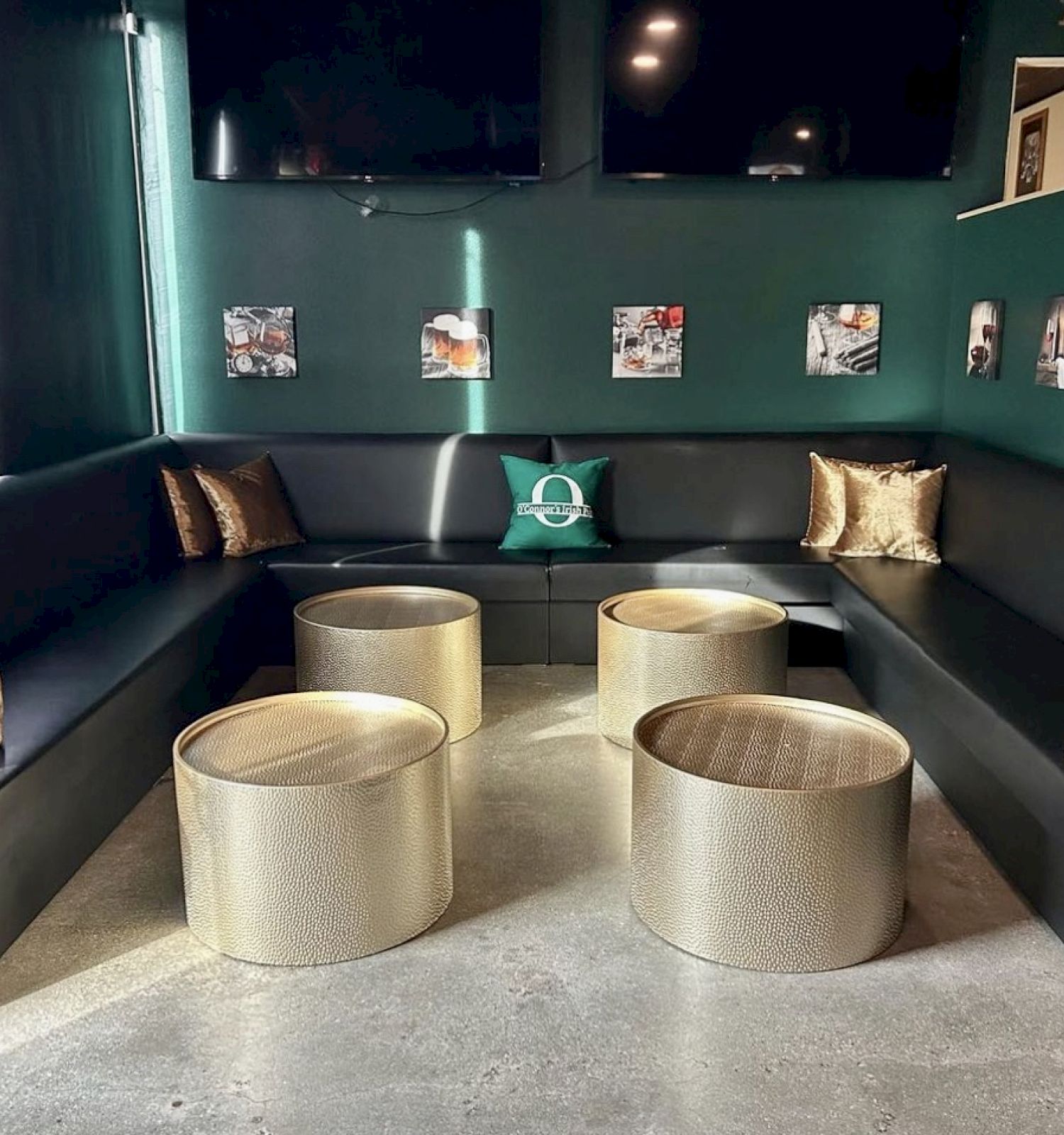 A modern lounge area with black cushioned seating, gold pillows, round tables, and artwork on green walls.