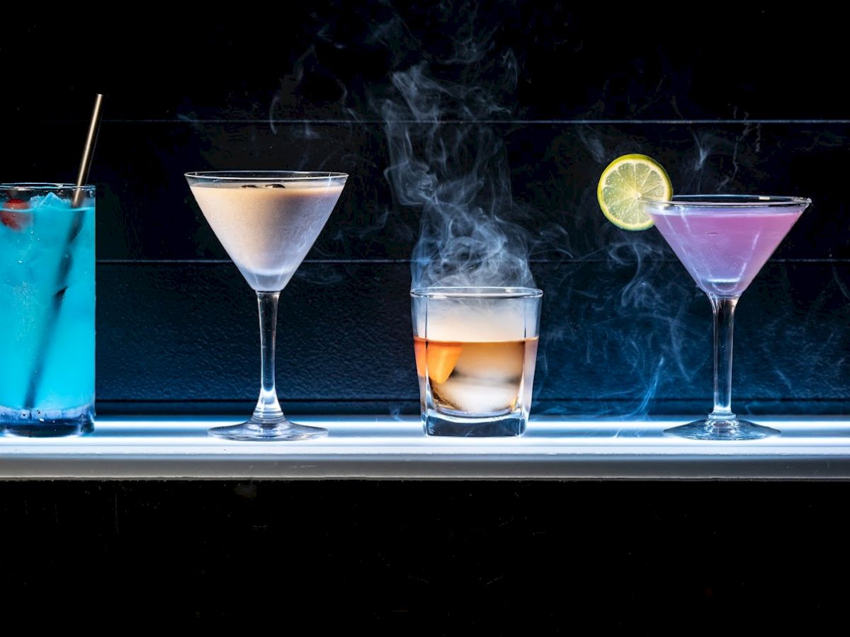 A row of four assorted cocktails: a blue-hued drink with a straw, two martini glasses with pink and creamy beverages, and a smoking glass.