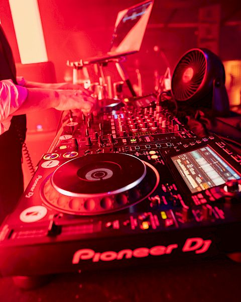 The image shows a DJ setup with turntables, a mixer, and a laptop, all illuminated by red lighting, featuring equipment branded 