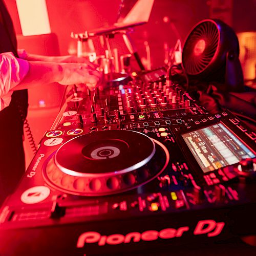 The image shows a DJ setup with turntables, a mixer, and a laptop, all illuminated by red lighting, featuring equipment branded 