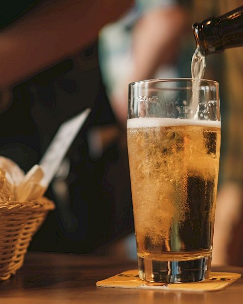 A glass of beer is being poured on a table with a coaster, next to a wicker basket containing bread or snacks.