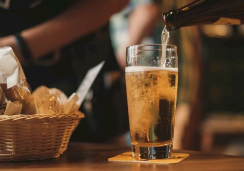 A glass of beer is being poured on a table with a coaster, next to a wicker basket containing bread or snacks.