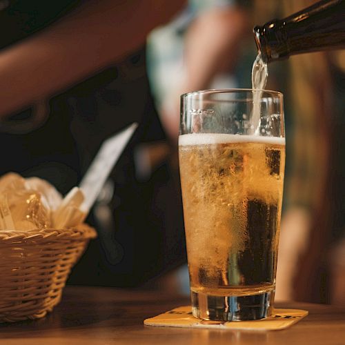 A glass of beer is being poured on a table with a coaster, next to a wicker basket containing bread or snacks.