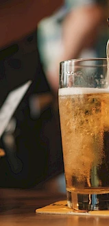 A glass of beer is being poured on a table, with a basket of bread on the side and a coaster underneath the glass.