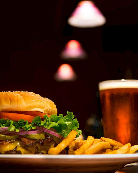 A burger with lettuce, tomato, onions, and cheese, accompanied by fries and a glass of beer on a table under dim lighting.