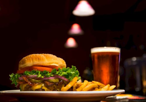 A burger with lettuce, tomato, onions, and cheese, accompanied by fries and a glass of beer on a table under dim lighting.