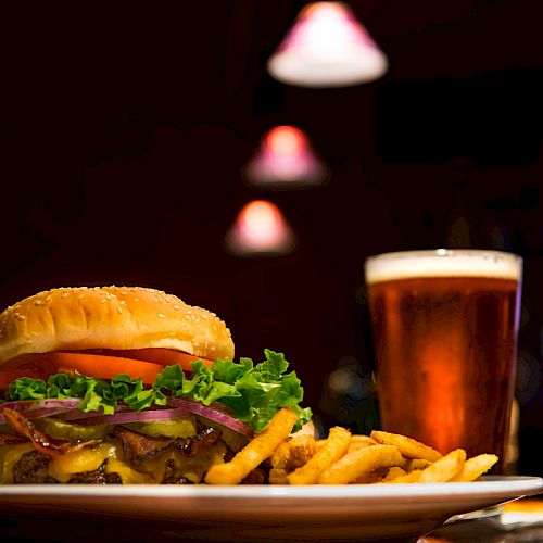 A burger with lettuce, tomato, onions, and cheese, accompanied by fries and a glass of beer on a table under dim lighting.