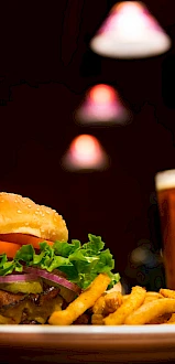 A burger with lettuce, tomato, and onions next to fries on a plate, accompanied by a glass of beer, under warm lighting at a dimly lit restaurant.