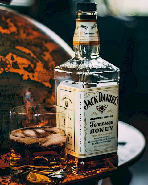 The image shows a bottle of Jack Daniel's Tennessee Honey whiskey and a glass with whiskey and ice cubes on a table.