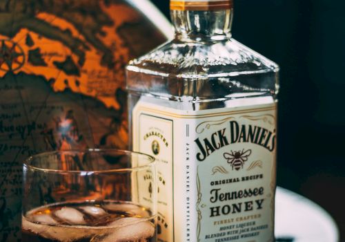 The image shows a bottle of Jack Daniel's Tennessee Honey whiskey and a glass with whiskey and ice cubes on a table.