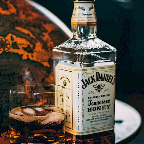 The image shows a bottle of Jack Daniel's Tennessee Honey whiskey and a glass with whiskey and ice cubes on a table.