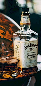 A bottle of Jack Daniel's Tennessee Honey whiskey and a glass with ice and whiskey sit on a wooden surface with a decorative background.