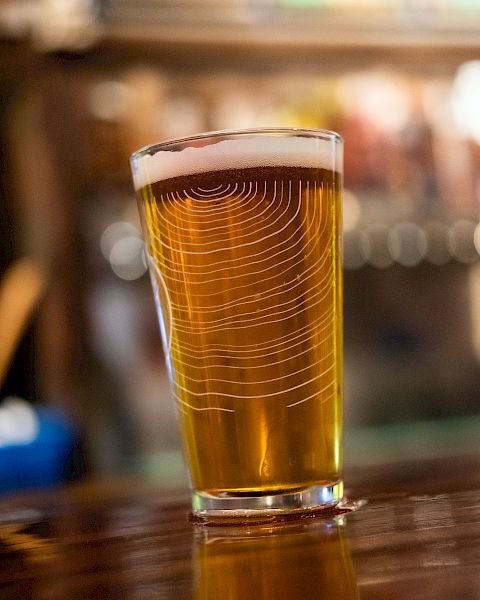 The image shows a pint of beer with a frothy head placed on a wooden surface, with a blurred bar background.