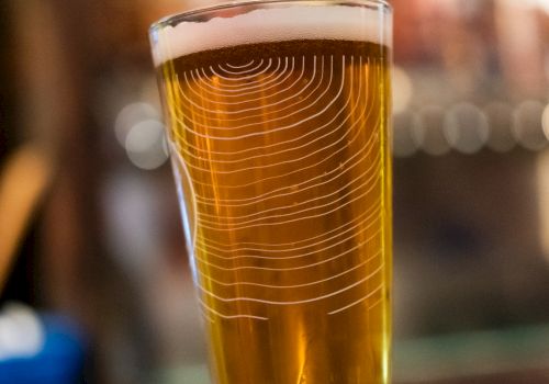 The image shows a pint of beer with a frothy head placed on a wooden surface, with a blurred bar background.