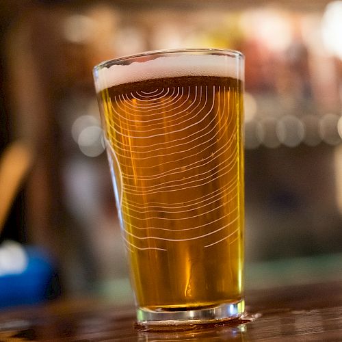 The image shows a pint of beer with a frothy head placed on a wooden surface, with a blurred bar background.