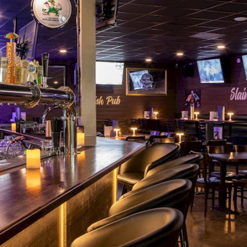 The image shows a cozy bar with a long counter, several bar stools, taps for beer, table seating, dim lighting, and multiple TVs on the wall.