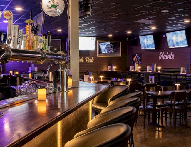 The image shows a cozy bar with a long counter, several bar stools, taps for beer, table seating, dim lighting, and multiple TVs on the wall.