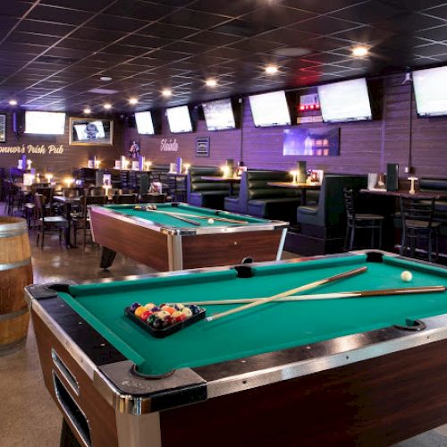 This image shows a bar with two pool tables, multiple TVs on the walls, booths for seating, and a wooden barrel with a bottle on top.