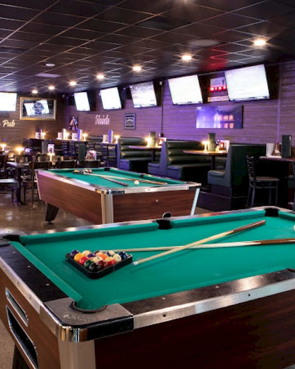 This image shows a bar with two pool tables, multiple TVs on the walls, booths for seating, and a wooden barrel with a bottle on top.